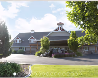Exterior view of a large building with a sloped roof, dormer windows, and a covered entrance surrounded by trees and landscaping under a partly cloudy sky.