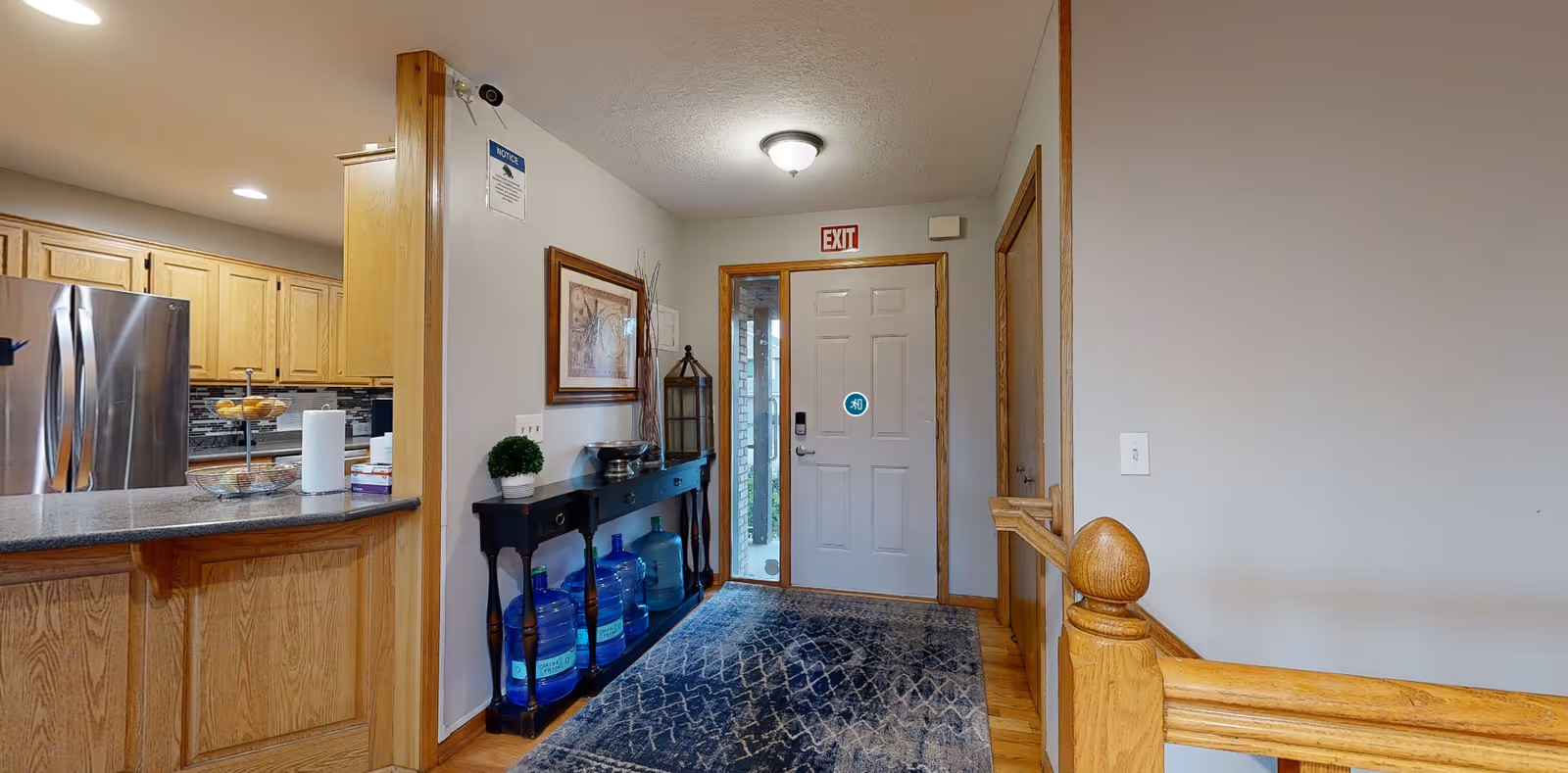 Interior entryway with a front door, rug, console table holding water jugs and decor, and a view into a kitchen with wooden cabinets.