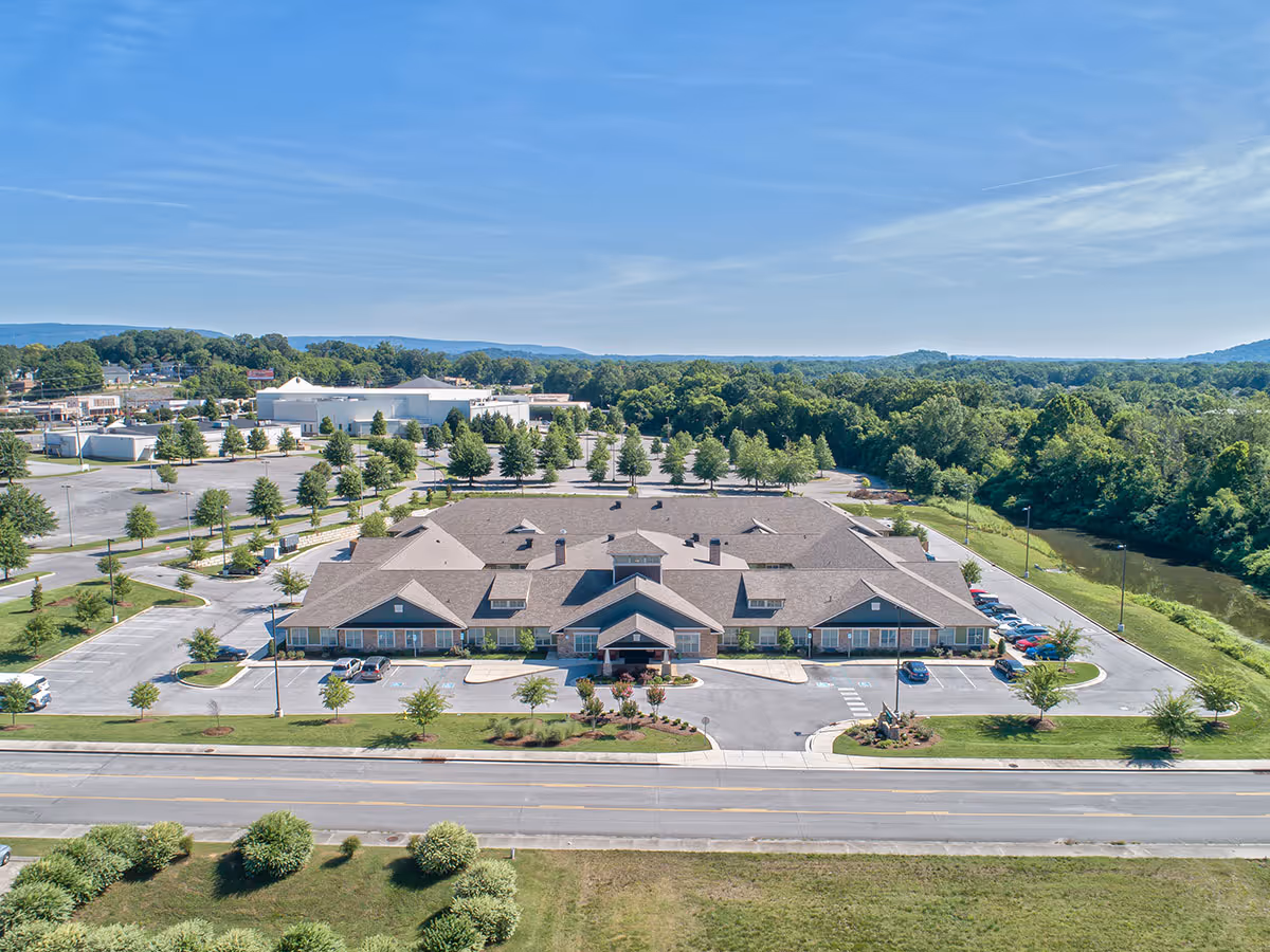 Aerial view of Dominion Senior Living of Hixson, a large single-story building with a gray roof surrounded by parking lots, trees, and greenery. The building is situated near a road with a river and forested area visible in the background under a clear blue sky.