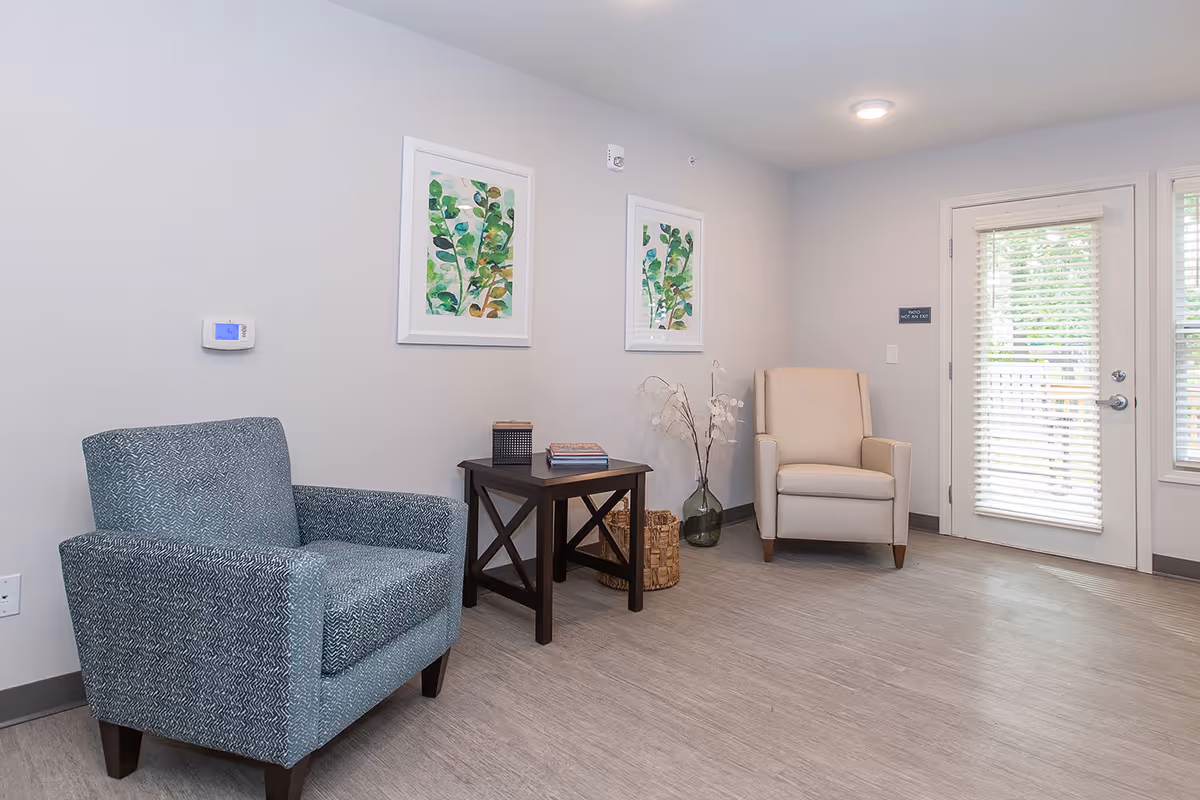 A small sitting area in a senior living facility with two armchairs, one blue patterned and one beige, separated by a dark wooden side table holding books and a decorative item. Two framed botanical prints hang on the light gray wall above the table. A glass vase with white flowers and a woven basket are placed on the floor next to the table. A door with white blinds leads outside, allowing natural light into the room.