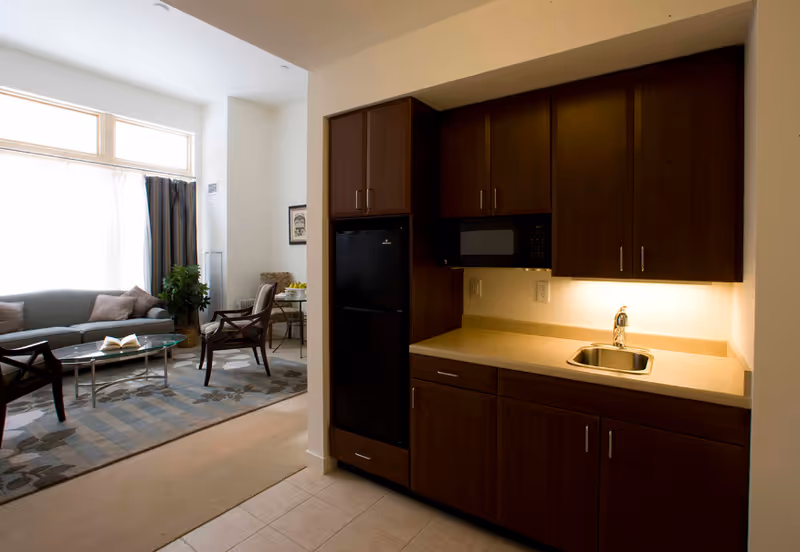 Interior view of a senior living facility showing a small kitchenette with dark wood cabinets, a sink, a microwave, and a refrigerator. Adjacent to the kitchenette is a living area with a sofa, two chairs, a glass coffee table, a patterned rug, and a window with curtains allowing natural light to enter.