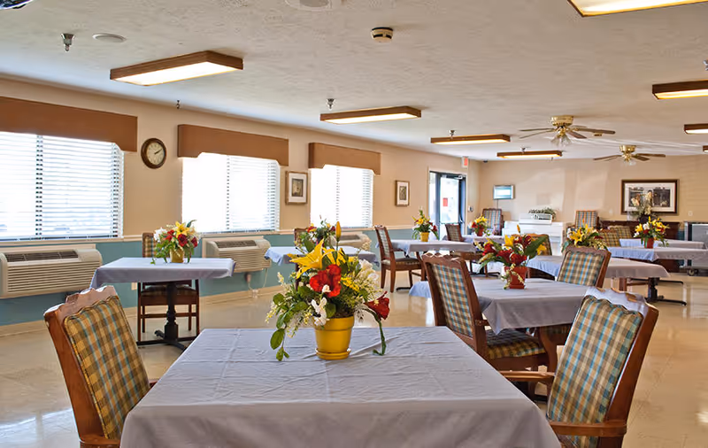 A bright dining room with multiple tables covered in light blue tablecloths, each adorned with colorful flower arrangements in yellow pots. The room has large windows with blinds, plaid upholstered chairs, ceiling fans, and framed pictures on the walls.