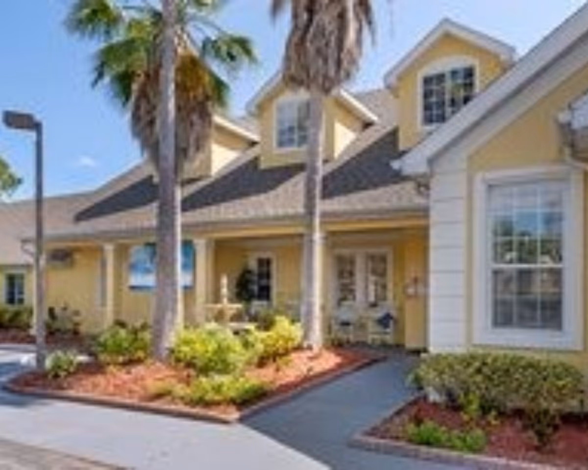 Yellow single-story building entrance with palm trees, landscaped beds and a walkway leading to double doors.