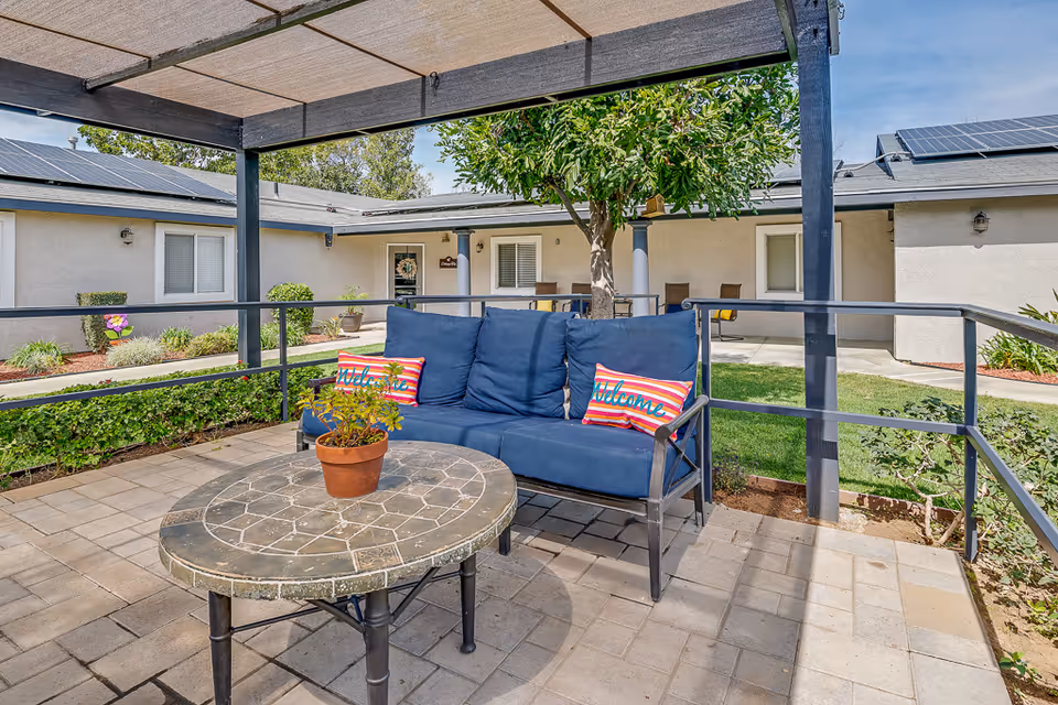 Outdoor patio area at Cottages at Riverside featuring a blue cushioned bench with two striped pillows that say 'Welcome', a round stone table with a potted plant, shaded by a pergola. Surrounding the patio are green bushes, a tree, and a building with windows and solar panels on the roof under a clear blue sky.