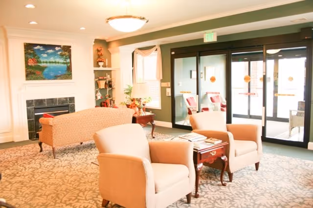 A bright and welcoming living room area in a retirement community featuring two beige armchairs with a small wooden table between them, a patterned carpet, a sofa near a fireplace with a landscape painting above it, and large glass doors leading outside.