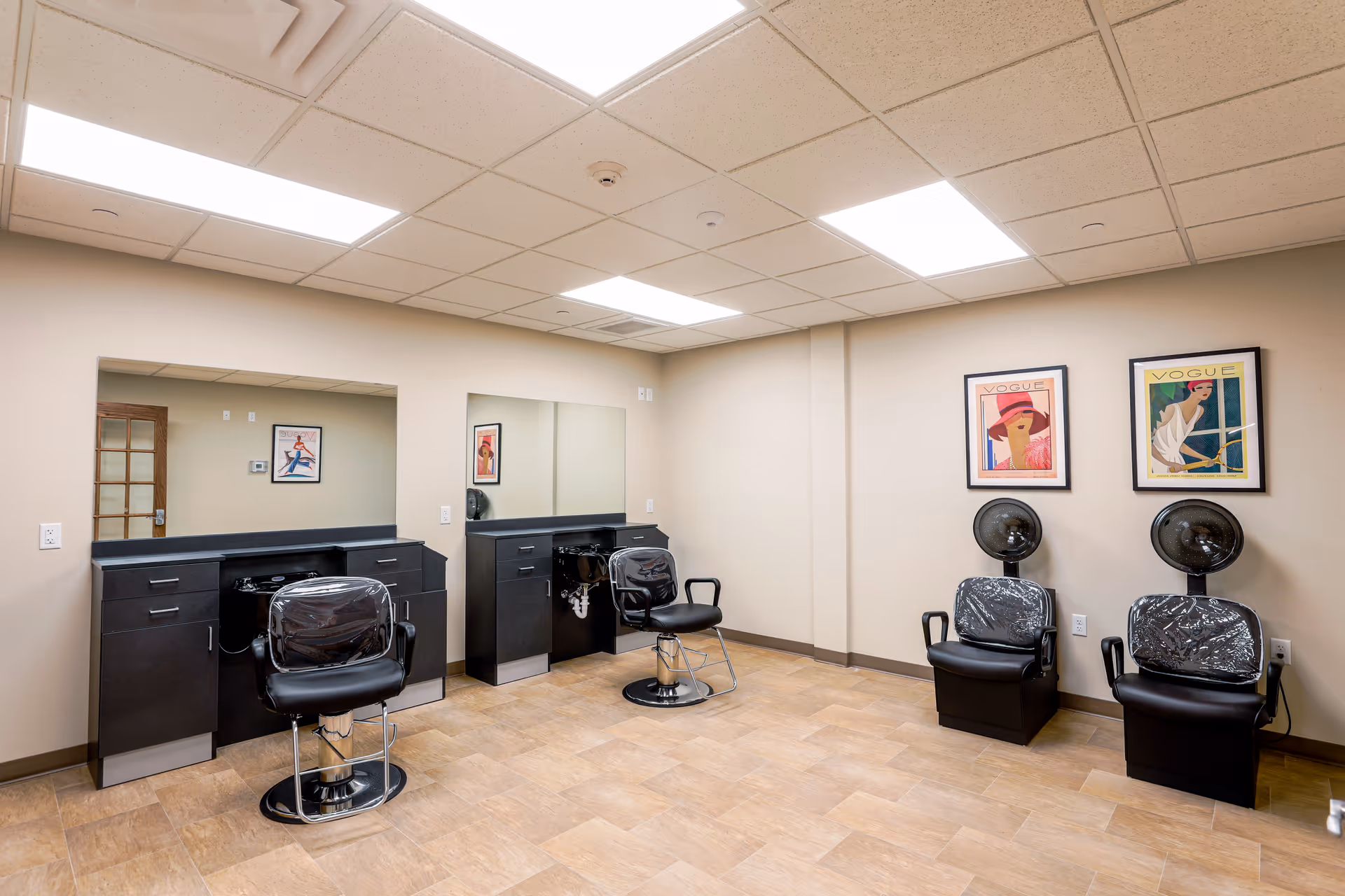 Interior view of a hair salon area in a senior living facility with two black salon chairs in front of large mirrors and two black hair drying chairs with hooded dryers against a beige wall. The room has a tiled floor and two framed Vogue magazine covers on the wall.