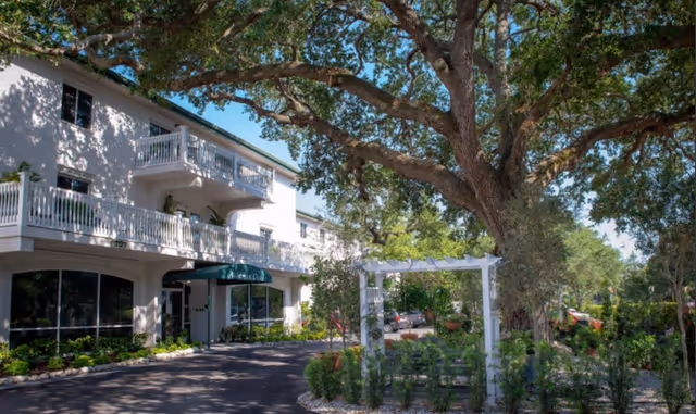 Exterior view of Alderman Oaks Retirement Residence showing a white multi-story building with balconies, surrounded by large trees and landscaped greenery including a white garden arbor and various plants.