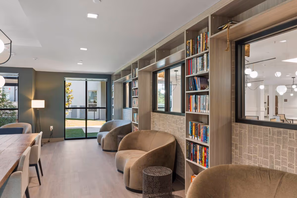 A modern, well-lit common area with a long wooden table and chairs on the left, and a row of plush armchairs along a wall with built-in bookshelves filled with books. Large windows provide natural light and a view of an outdoor courtyard.