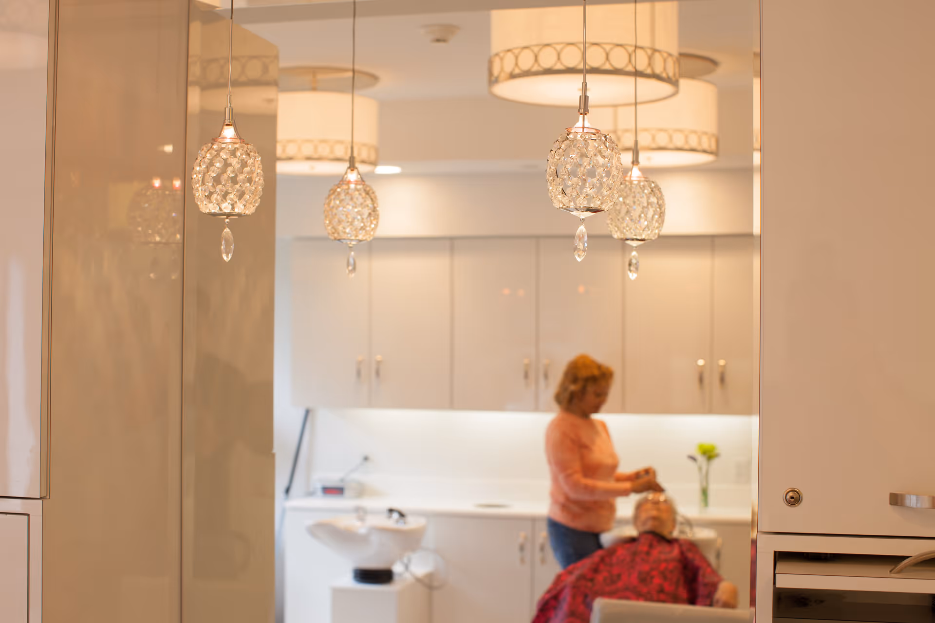 Interior view of a salon area with crystal pendant lights and a staff member washing a resident's hair at a sink.