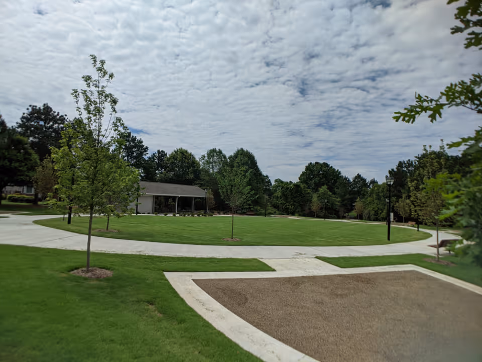 A landscaped outdoor area with a circular paved walkway surrounding a grassy lawn, young trees planted along the path, a small pavilion or shelter in the background, and a partly cloudy sky overhead.
