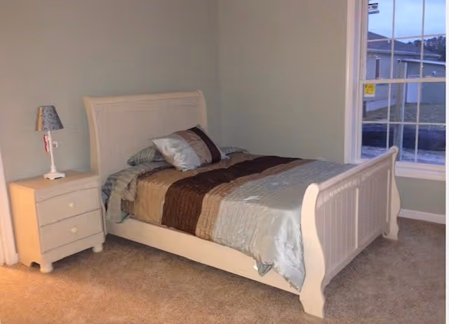 Small bedroom with a white wooden bed made with brown and light blue bedding, a bedside table with a lamp, and a window looking outside.