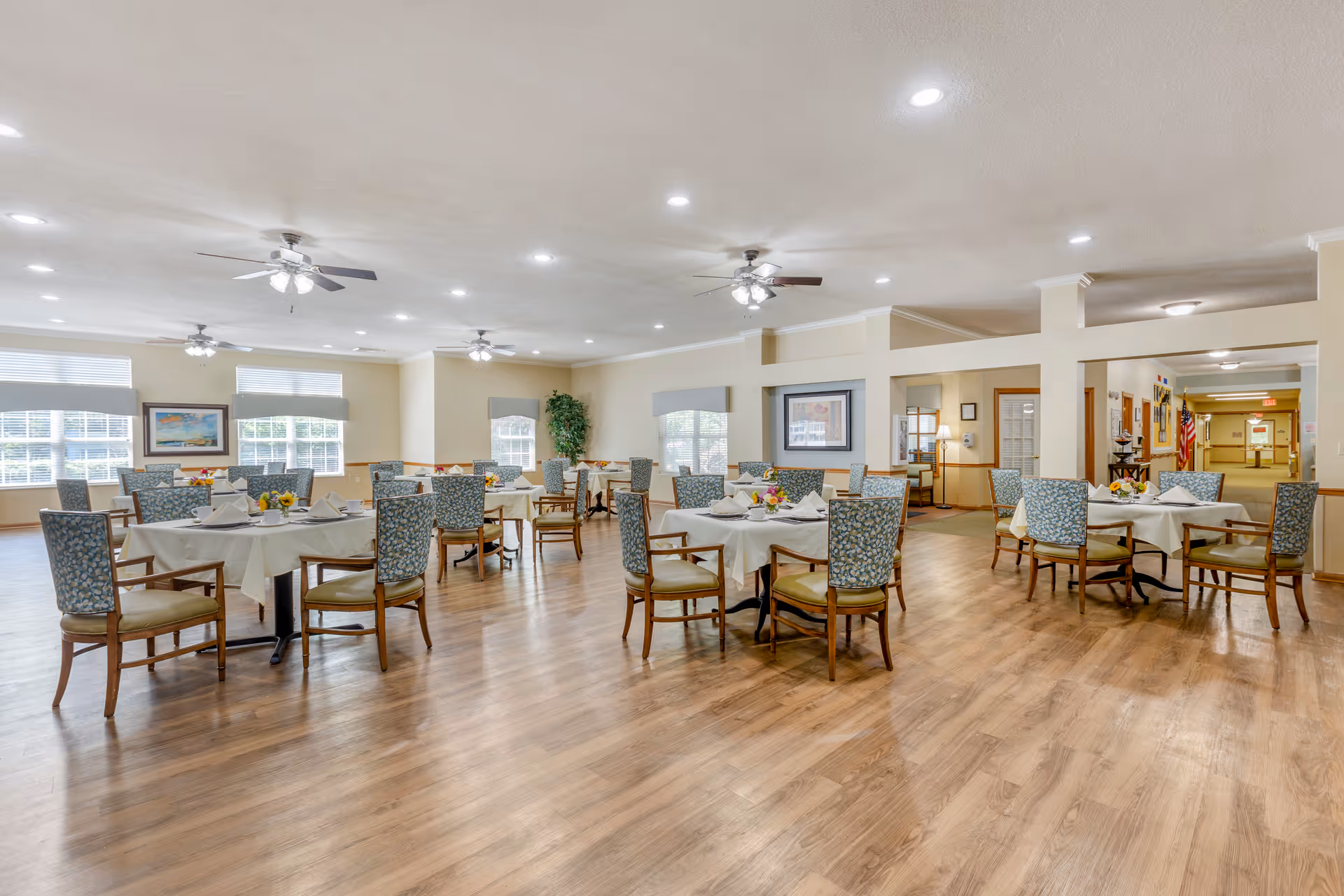 Spacious dining room with round tables set for meals and upholstered chairs on wood flooring under ceiling fans and recessed lights.