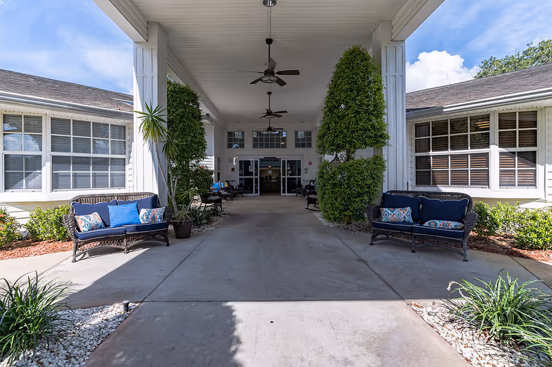 Covered entrance area of a senior living facility with two wicker benches with blue cushions and decorative pillows on either side. There are potted plants and trimmed bushes along the walkway leading to glass double doors. Ceiling fans are mounted on the white ceiling above the entrance.