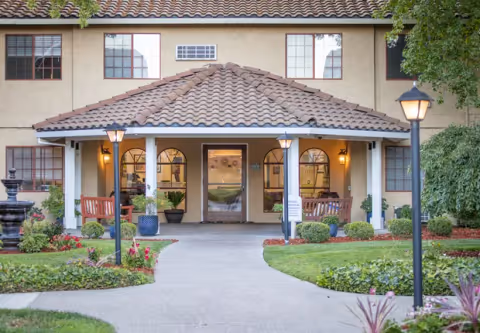 Front entrance of a senior living facility with a tiled roof, arched windows, and a pathway leading to the door. There are benches, potted plants, lamp posts, and well-maintained landscaping with green grass and flowers.