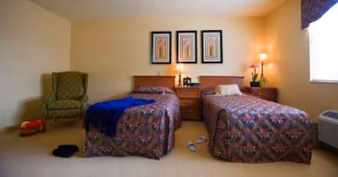 A bedroom with two twin beds covered in patterned bedspreads, a nightstand with a lamp and a coffee maker between the beds, a green upholstered armchair in the corner, slippers on the floor, and a window with a valance letting in natural light.