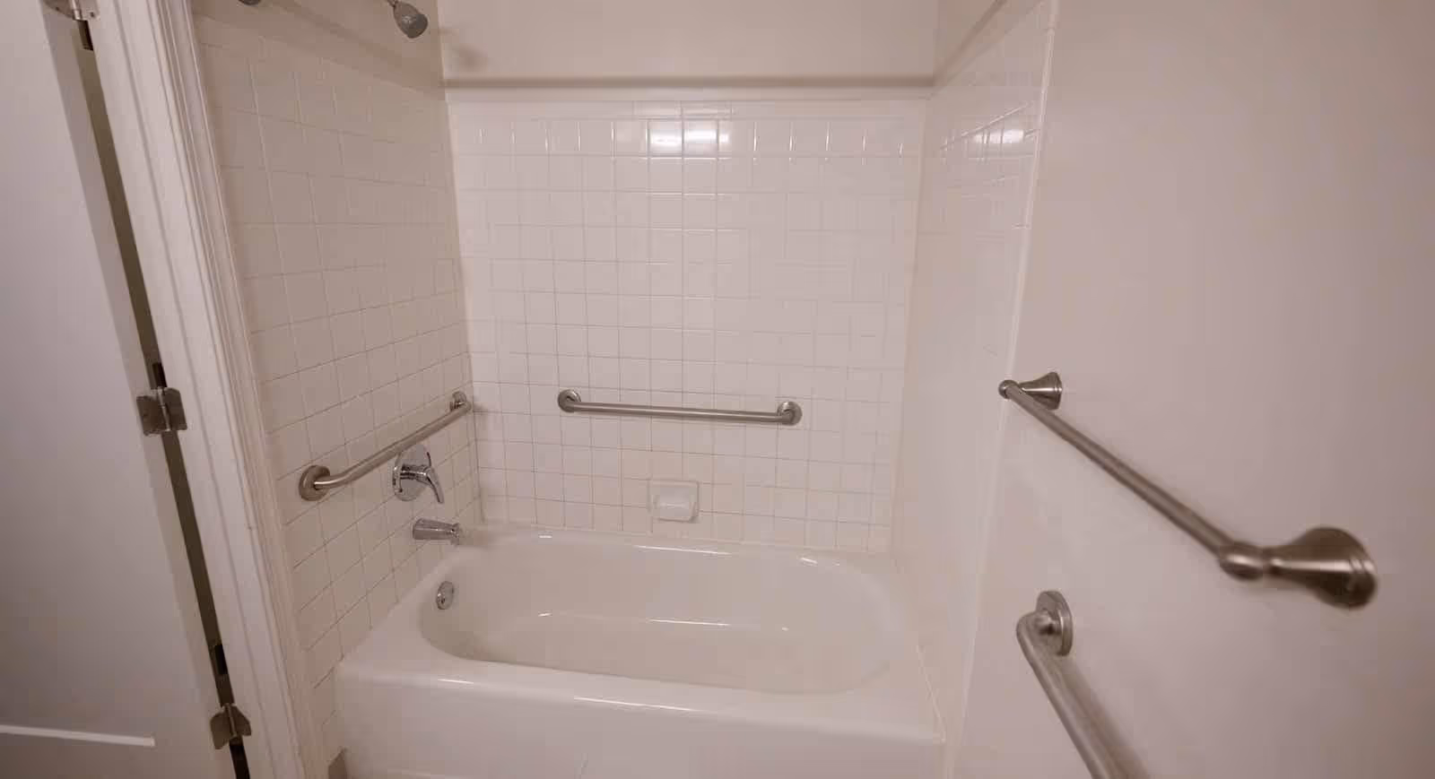 A white bathtub with tiled walls surrounding it. There are three metal grab bars installed on the walls around the bathtub for safety. A showerhead is mounted on the wall above the tub, and a towel bar is visible on the right wall.