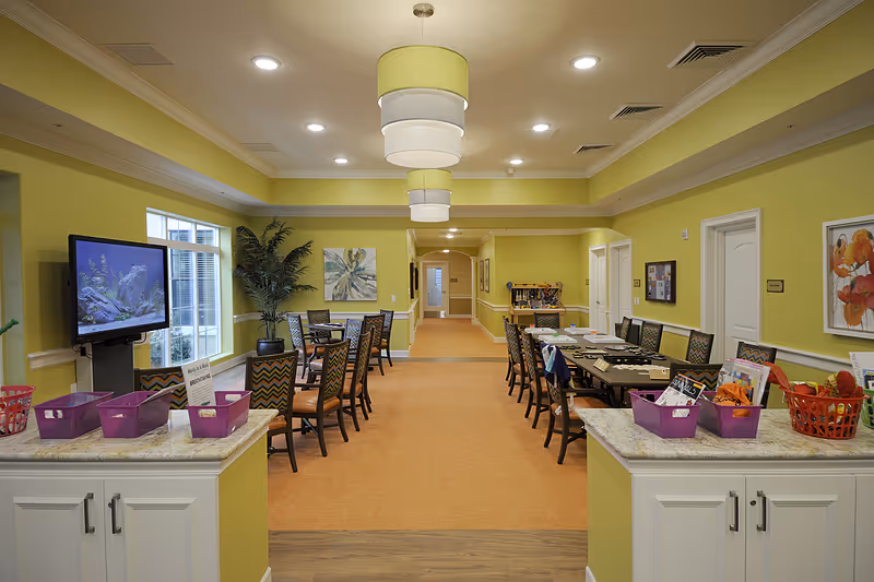 Bright communal dining and activity room with long tables, chairs, pendant lights, and craft baskets in a senior living facility.