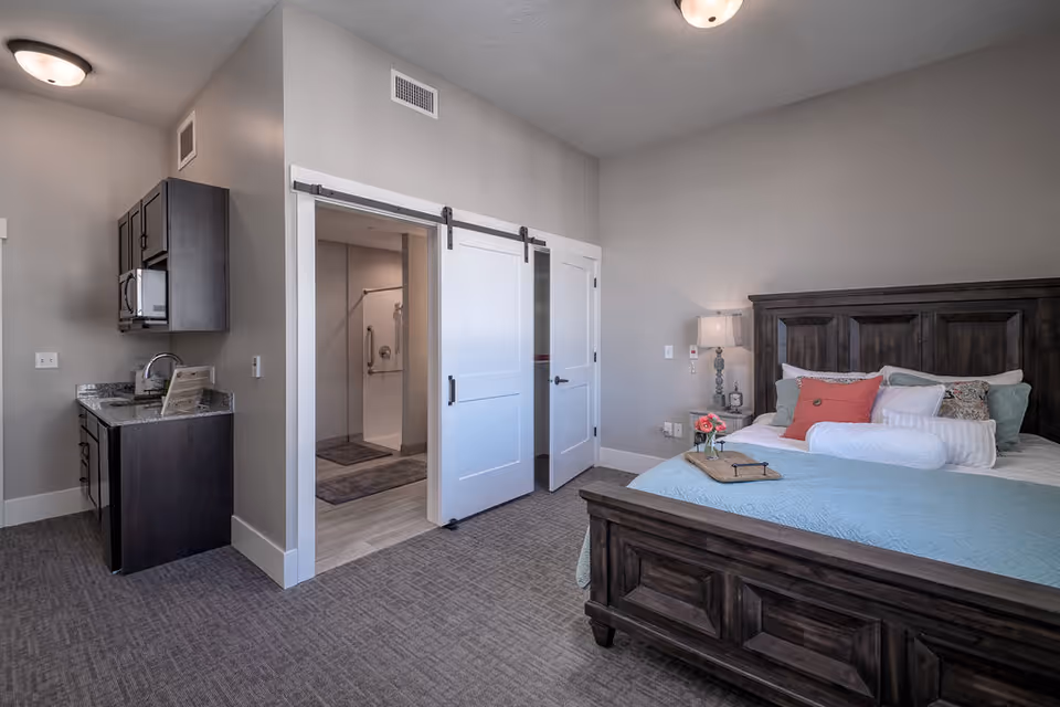A senior living facility bedroom with a dark wooden bed frame, a bed made with blue and white bedding and multiple pillows, a bedside table with a lamp and flowers, a kitchenette with dark cabinets and a granite countertop, and an open doorway leading to a bathroom with a walk-in shower.