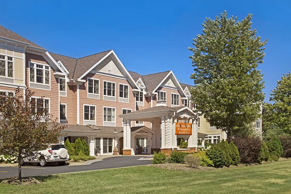Exterior view of a multi-story senior living facility building with a covered entrance, surrounded by trees and landscaping under a clear blue sky.