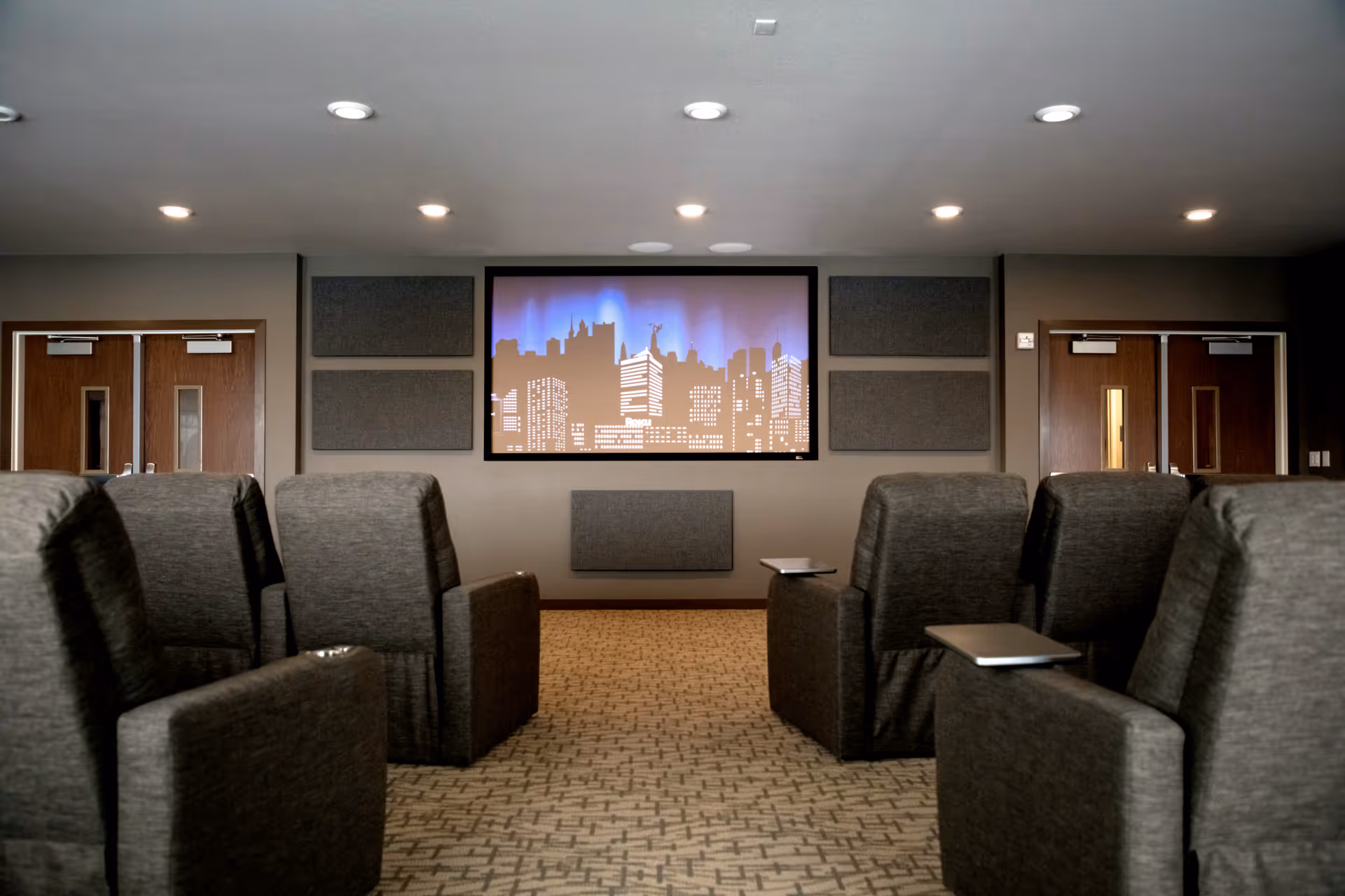 A small theater room with rows of gray upholstered recliner chairs facing a large screen displaying a city skyline graphic. The room has a patterned carpet, recessed ceiling lights, and double wooden doors on both sides of the screen.