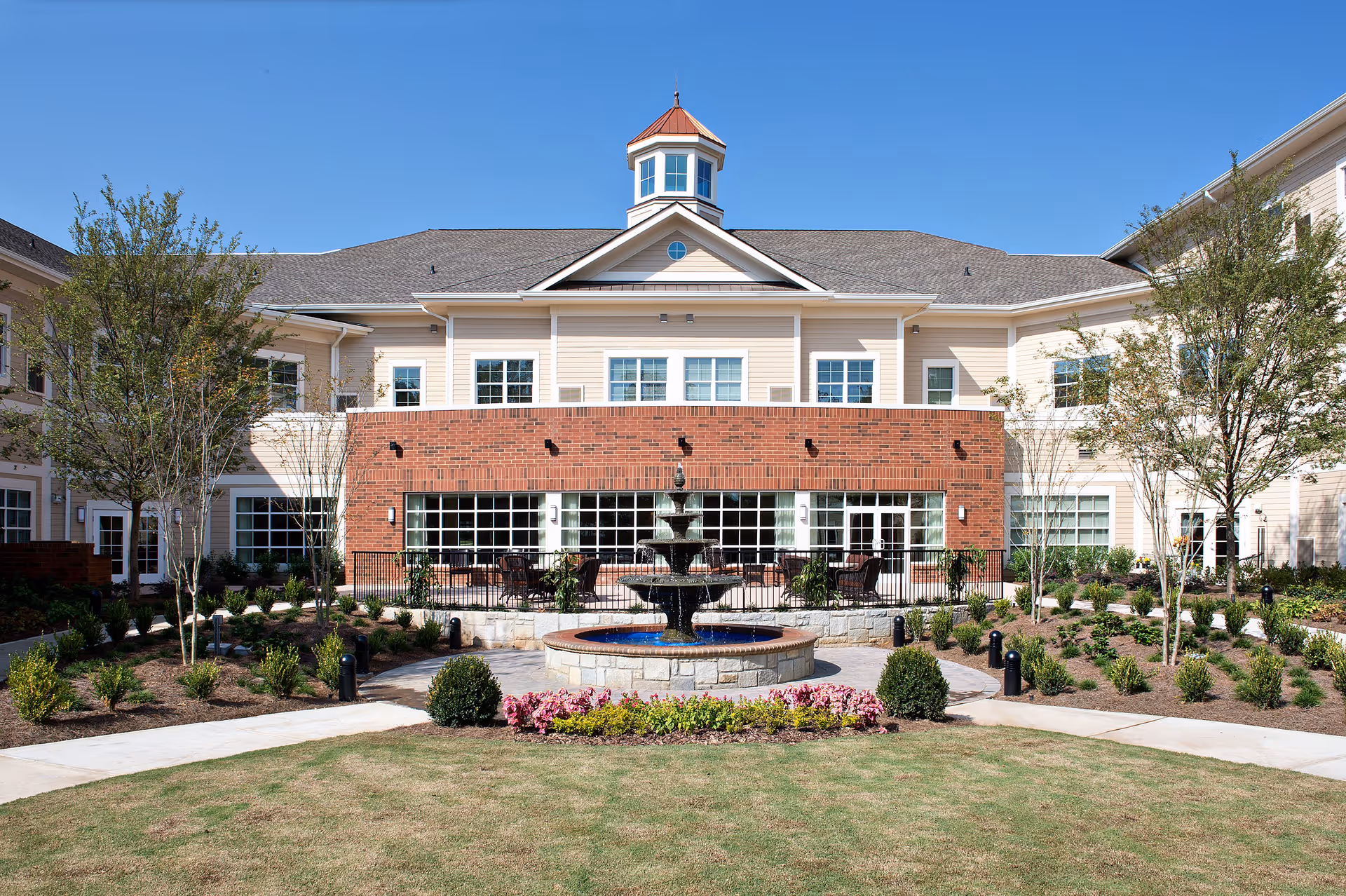 Exterior view of Sterling Estates of East Cobb Retirement Community building with a central water fountain, landscaped garden, and clear blue sky.