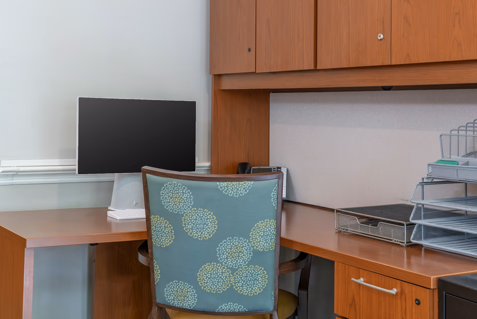 A wooden desk workspace with a monitor, patterned chair, metal document trays, and overhead cabinets in an office nook.
