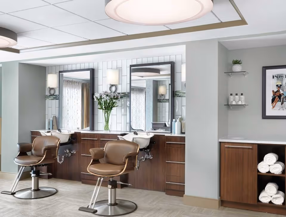 Interior view of a salon area in a senior living community featuring two brown salon chairs in front of a counter with sinks and large mirrors. The walls are light gray with white tiled backsplash, and there are shelves with rolled white towels and bottles. A framed picture and a small potted plant are on the wall.
