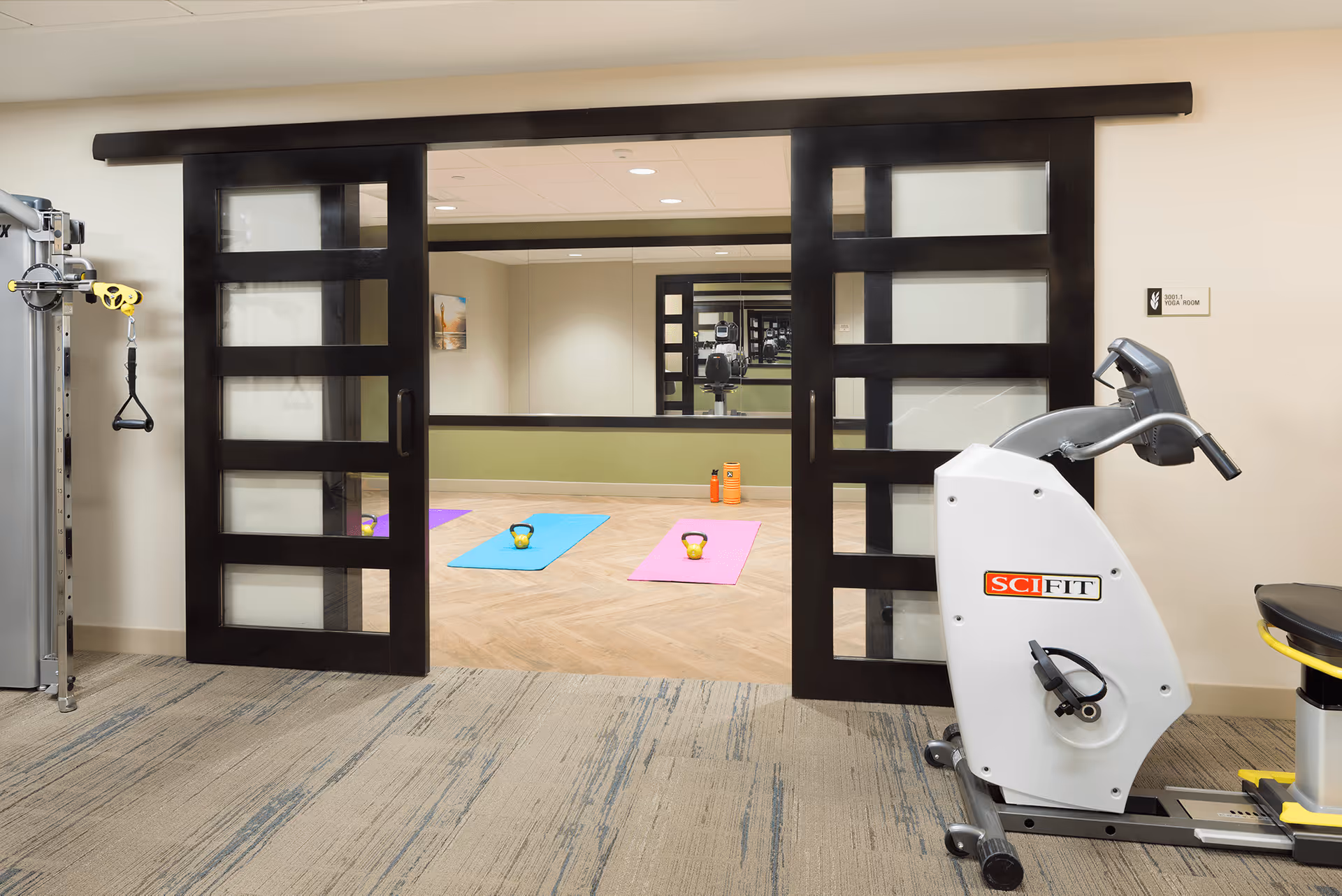 View of a fitness room in a senior living facility with exercise equipment including a SciFit stationary bike and a cable machine. Through large sliding black-framed glass doors, a yoga room is visible with two yoga mats, each with a kettlebell placed on top, and two orange water bottles against the far wall. The yoga room has a large mirror on the back wall and light wood flooring.