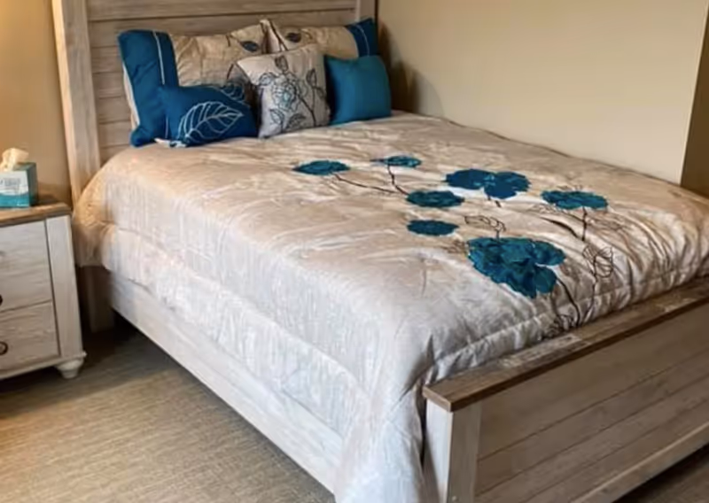 A neatly made bed with a light-colored wooden frame and headboard, covered with a beige comforter featuring blue floral designs. The bed has several decorative pillows in shades of blue and beige. Next to the bed is a matching wooden nightstand with a box of tissues on it.