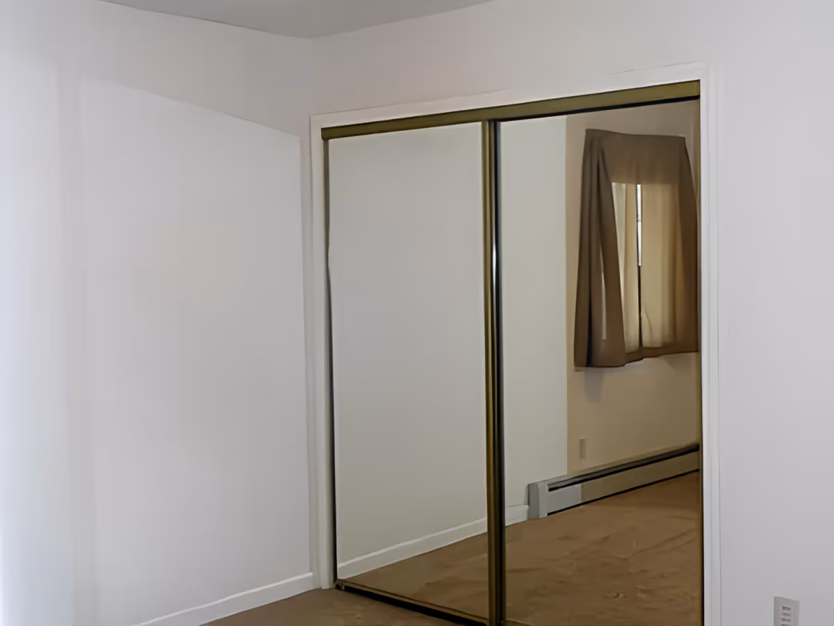 Empty room with beige carpet and white walls featuring a large closet with sliding mirrored doors reflecting a window with brown curtains and a baseboard heater.
