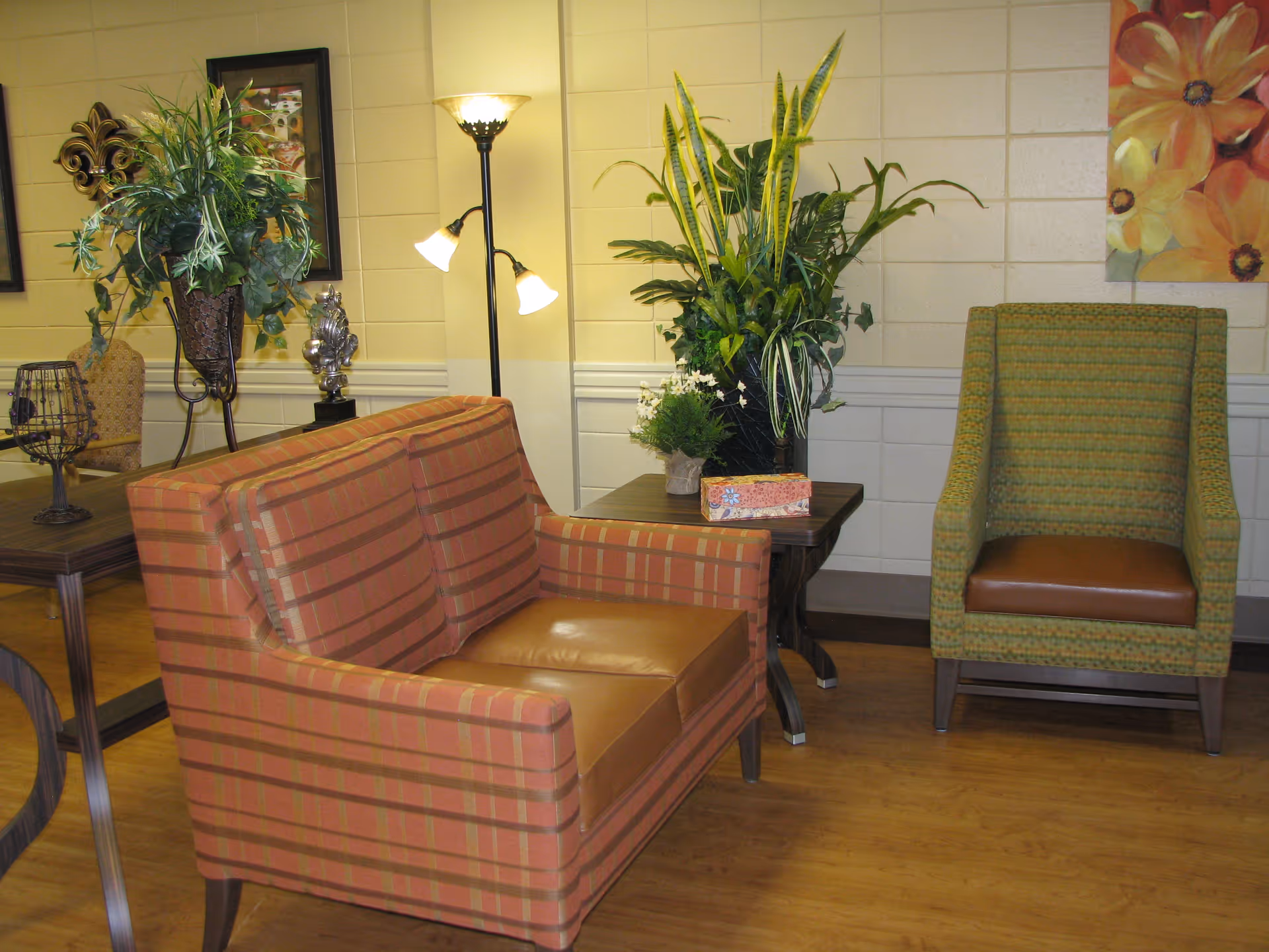 A cozy seating area in a community care center with a striped orange loveseat and a green patterned armchair. There are two side tables with decorative plants and a floor lamp with three lights. The walls are light-colored with framed artwork and floral paintings.