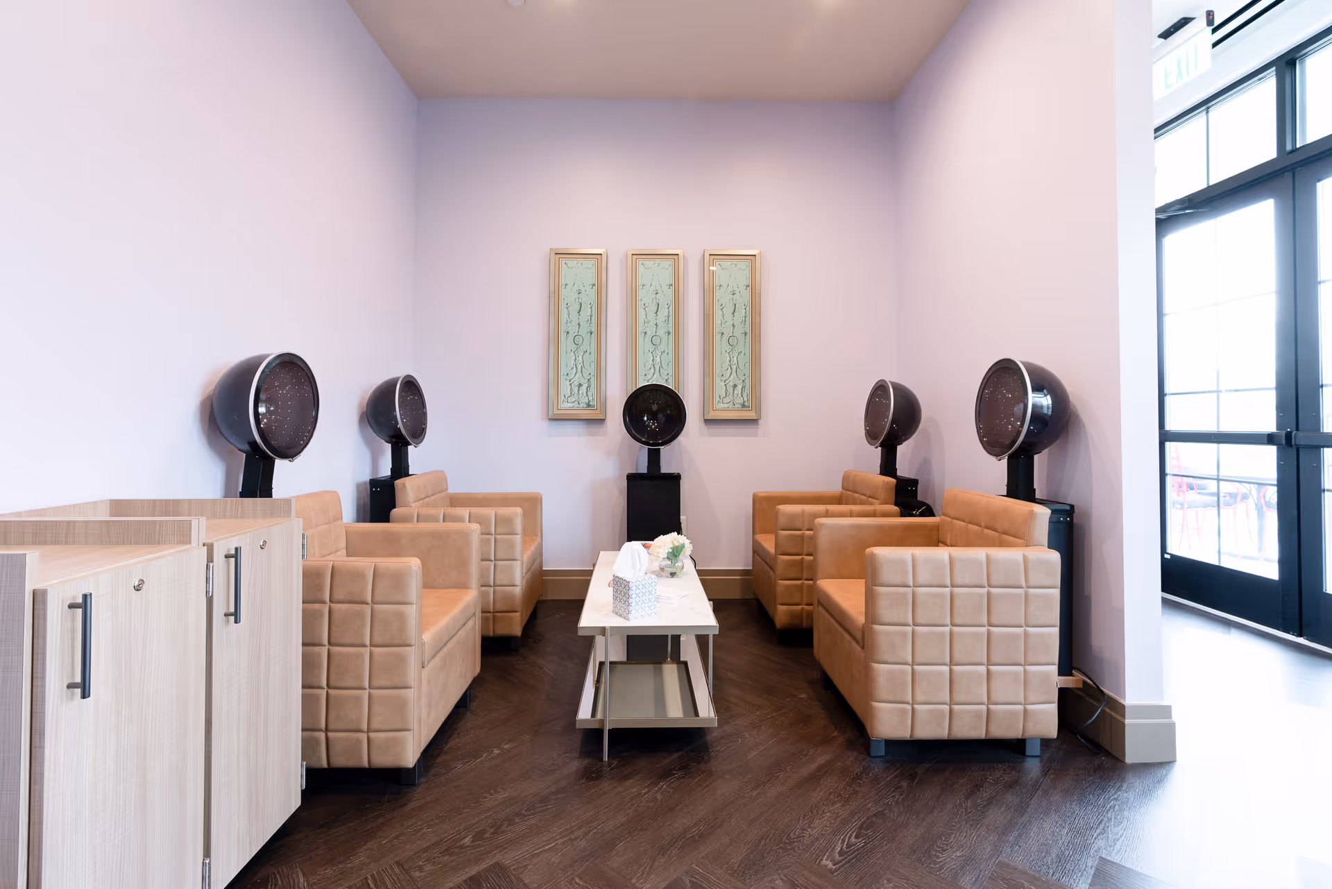 A salon area with four beige leather chairs arranged in two rows facing each other, each chair paired with a black hair dryer hood. A small white table with a tissue box and flower arrangement is placed in the center. The walls are light lavender, and there are three decorative framed panels on the back wall. To the right, there is a glass door letting in natural light.
