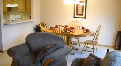 A cozy dining area with a wooden oval table set with red bowls and small plants, surrounded by four wooden chairs. In the foreground, there are two cushioned armchairs with patterned pillows. The dining area is adjacent to a kitchen with wooden cabinets and a white stove visible through a pass-through window. A framed picture hangs on the wall above the dining table, and a hanging light fixture illuminates the space.