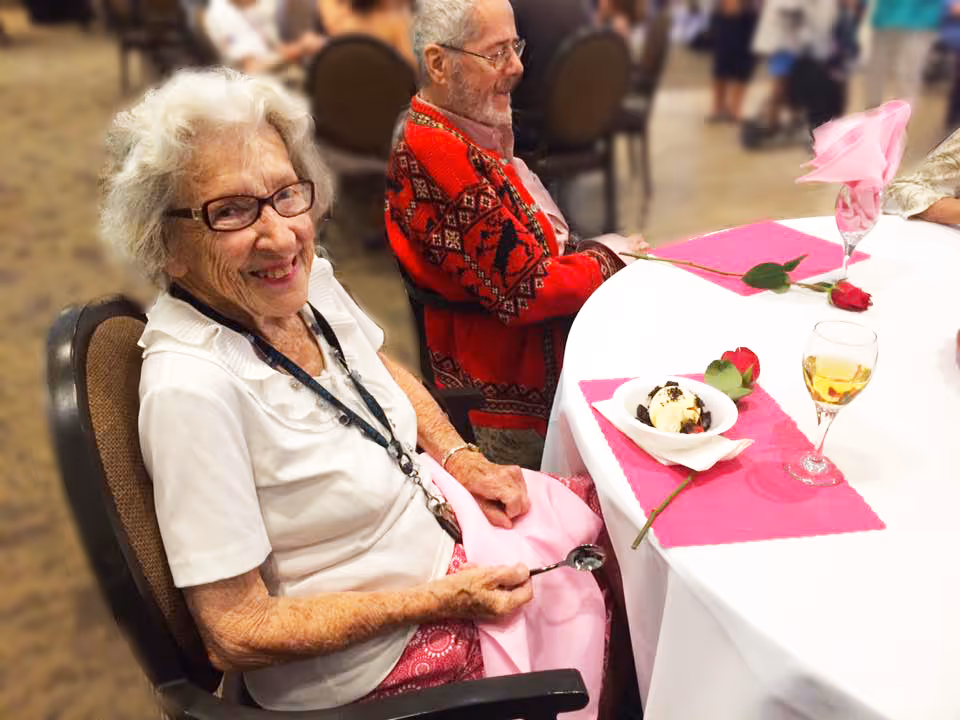 An elderly woman with white hair and glasses smiling while seated at a round table with a white tablecloth and pink placemats. She is holding a spoon and has a dessert bowl in front of her. Another elderly person wearing a red patterned sweater is seated next to her. The setting appears to be a dining area with other people and tables in the background.