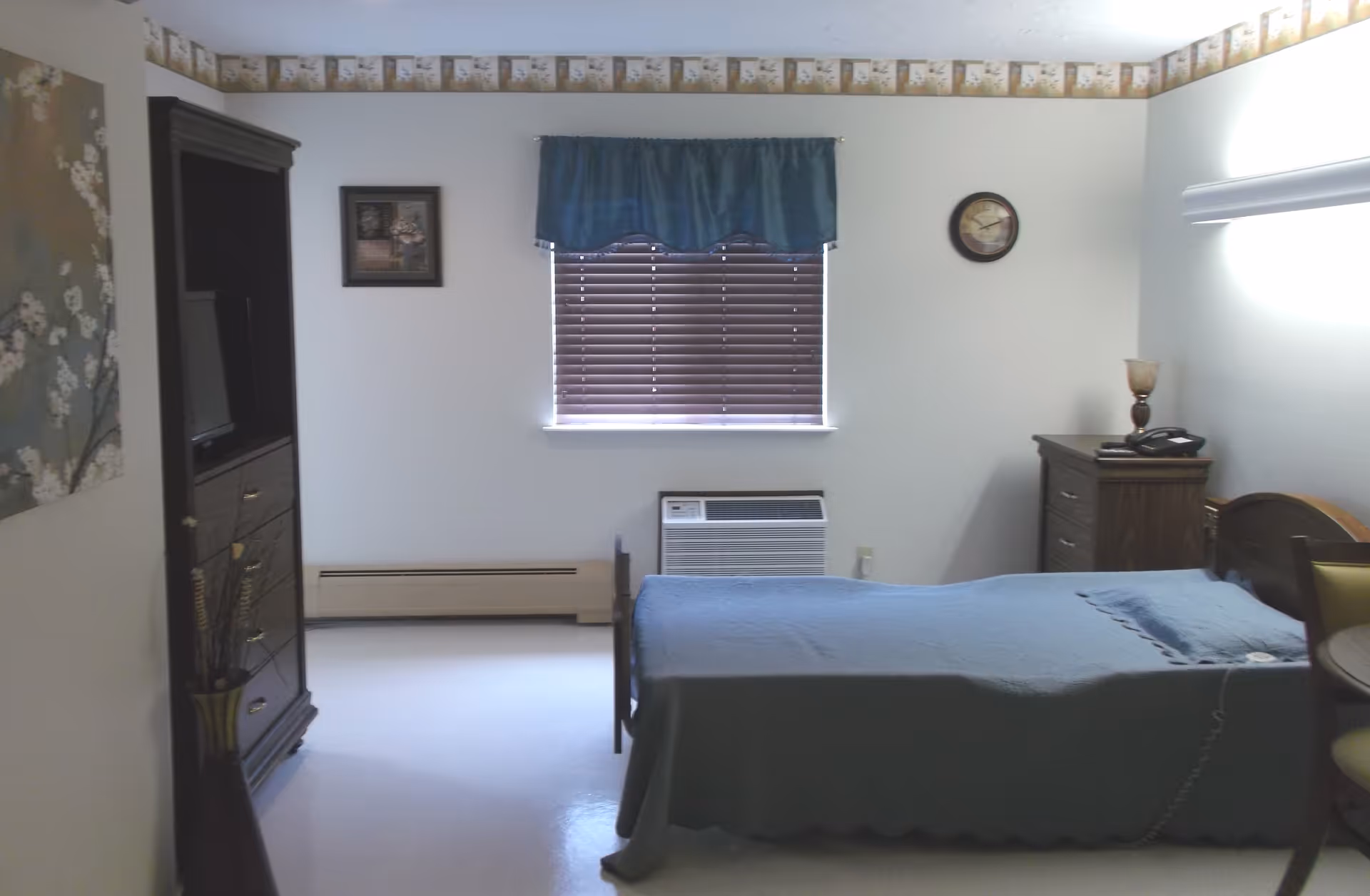 A simple bedroom with a single bed covered in a blue blanket, a wooden nightstand with a lamp and telephone, a window with closed blinds and a blue valance, a wall clock, a wooden cabinet with a TV, and a vase with decorative branches.