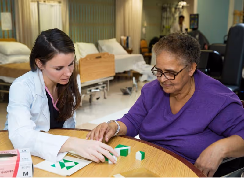 A healthcare worker in a white coat is sitting at a round table with an elderly woman wearing glasses and a purple top. They are engaged in an activity involving green and white blocks. In the background, there are hospital beds and medical equipment, indicating a care facility setting.