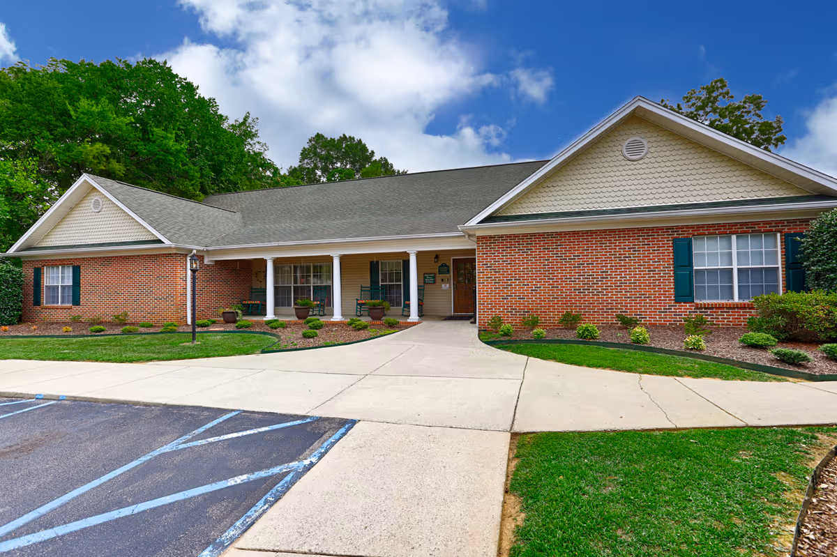 Exterior view of a single-story brick building with a covered porch supported by white columns. The porch has green rocking chairs and potted plants. The building is surrounded by neatly maintained landscaping with green grass, shrubs, and trees in the background under a partly cloudy blue sky. A paved walkway and parking area with handicap markings are visible in the foreground.