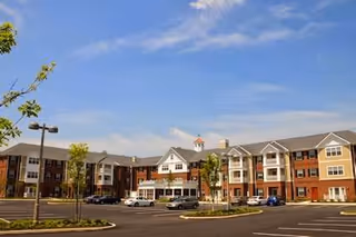 Front exterior of a three-story senior living building with a paved parking lot, landscaped islands, and a few cars under a blue sky.