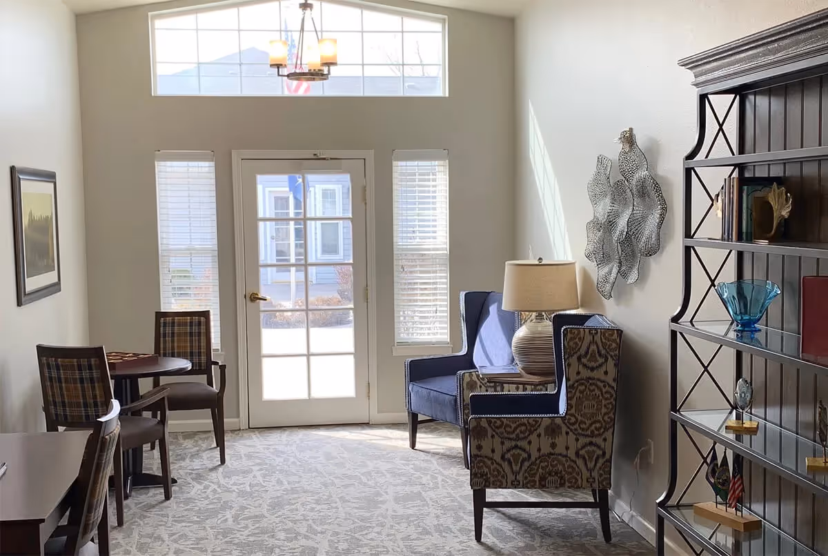 Bright communal seating area with upholstered chairs, a side table and lamp, bookshelf and a glass-paneled door to the outside.