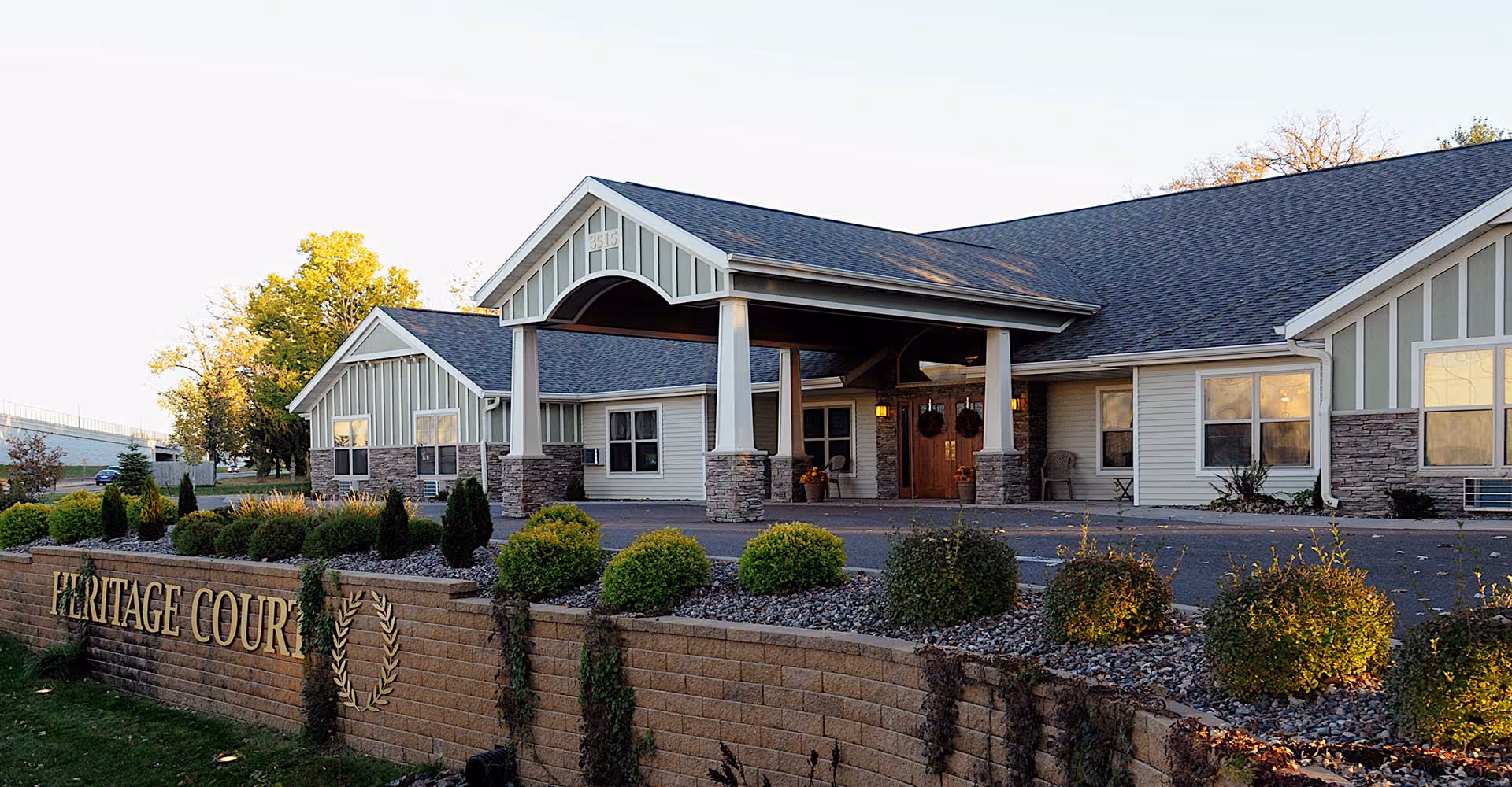 Single-story senior living facility entrance with a covered porte-cochere and a 'Heritage Court' sign on a low stone wall.