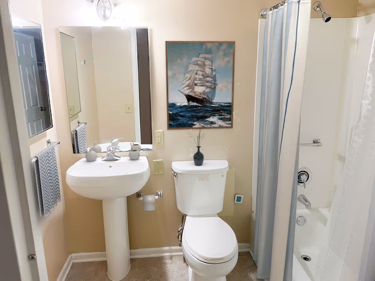 A small bathroom with a white pedestal sink, a toilet with a small black vase on top, a shower with a blue and white striped curtain, and a framed picture of a sailing ship on the wall above the toilet. There is a mirror above the sink and a towel hanging on a rack to the left.