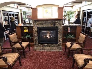Cozy lounge with a marble fireplace at center, upholstered chairs, bookshelves, and a reception desk with two staff members.