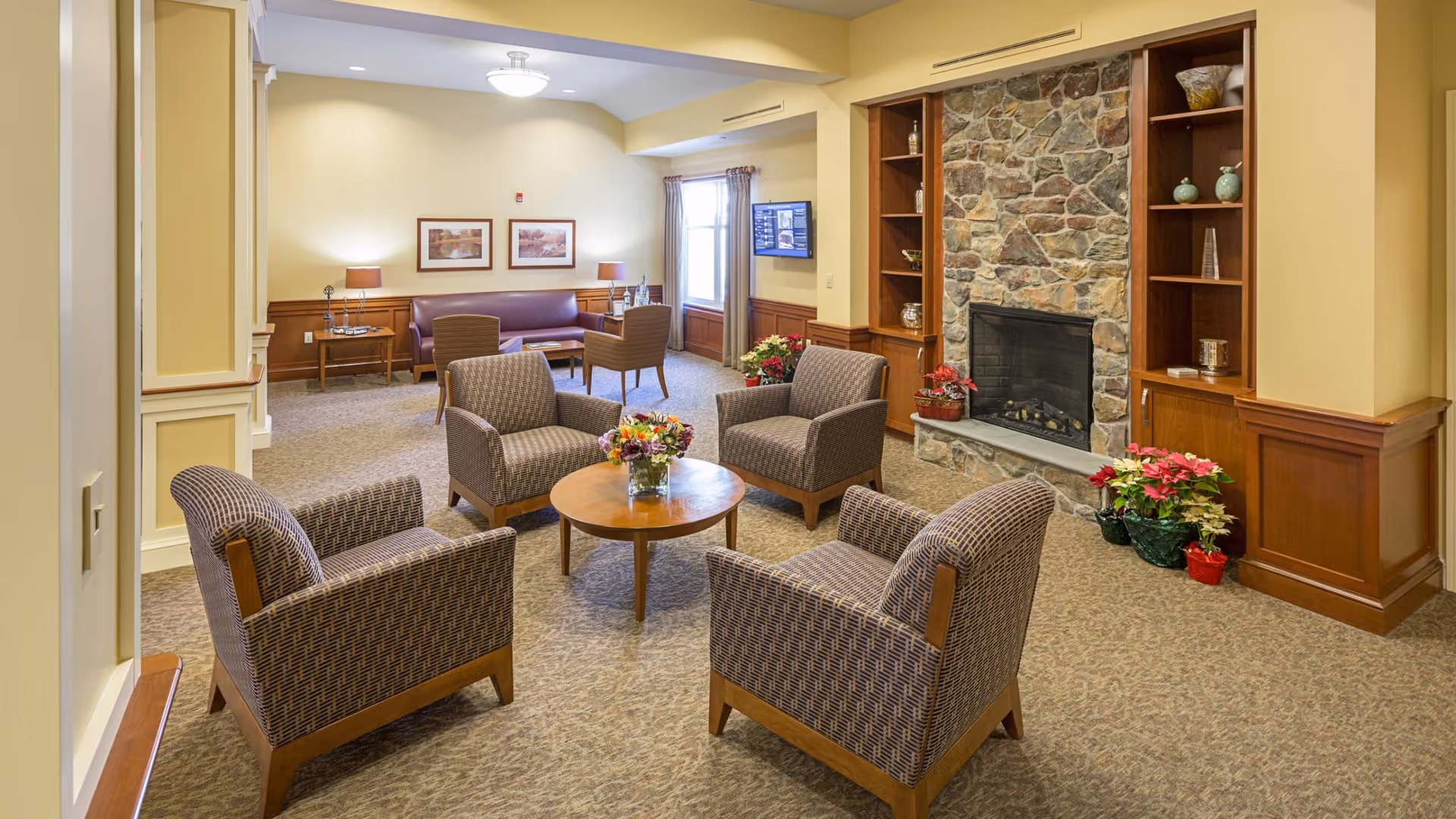 A cozy living room area with four patterned armchairs arranged around a round wooden coffee table with a flower arrangement. In the background, there is a stone fireplace flanked by wooden shelves with decorative items and potted plants. A purple sofa and two additional chairs are positioned near a window with curtains, and a wall-mounted TV is visible. The room has beige walls, carpeted floors, and warm lighting.