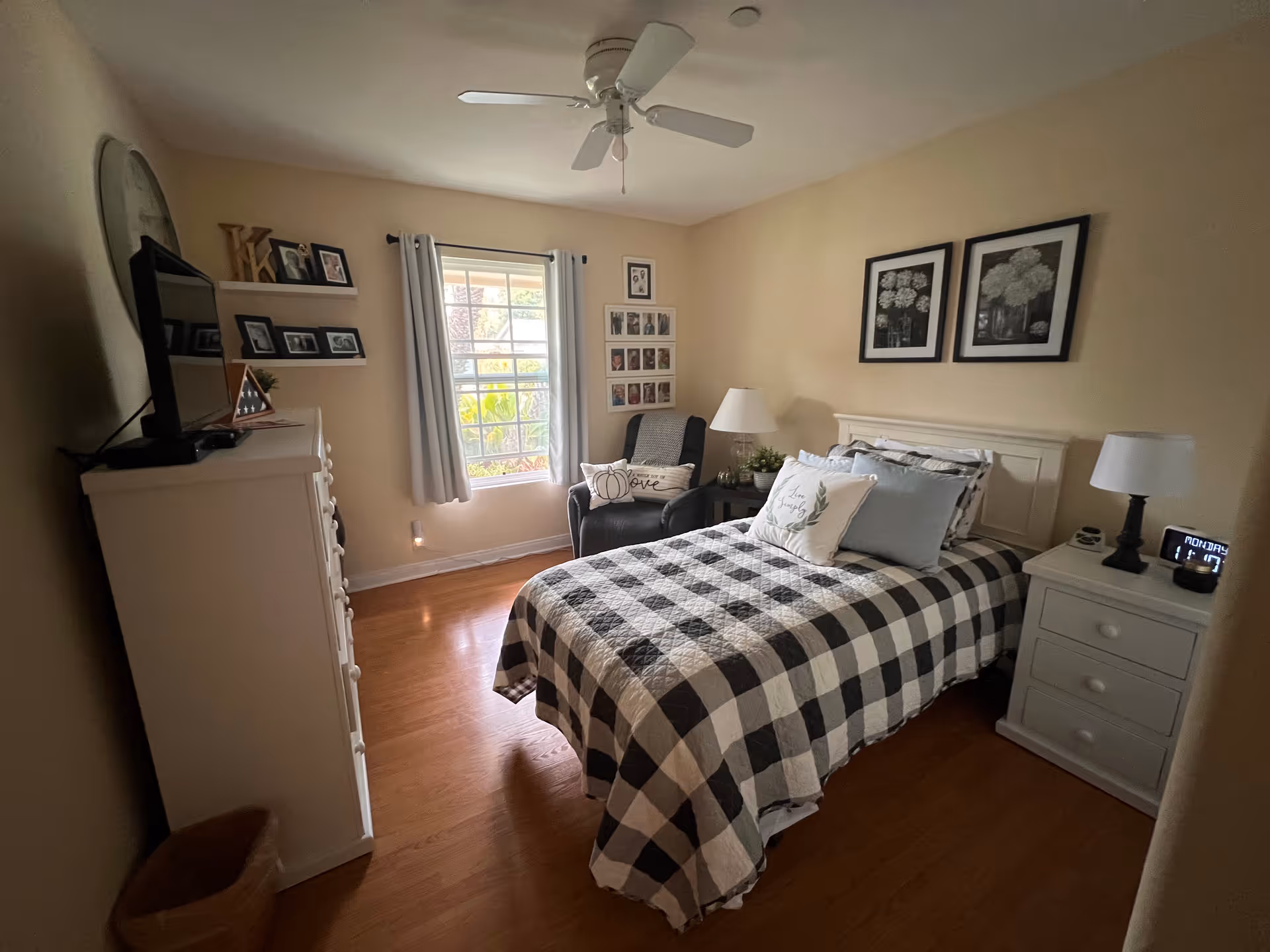 A cozy bedroom with a single bed covered in a black and white checkered quilt. The room has light beige walls, wooden flooring, and a white ceiling fan. There is a white dresser with a TV on top on the left side, and a white nightstand with a lamp and a digital clock on the right side of the bed. A comfortable armchair with a pillow is positioned near a window with white curtains. The walls are decorated with framed pictures and artwork.