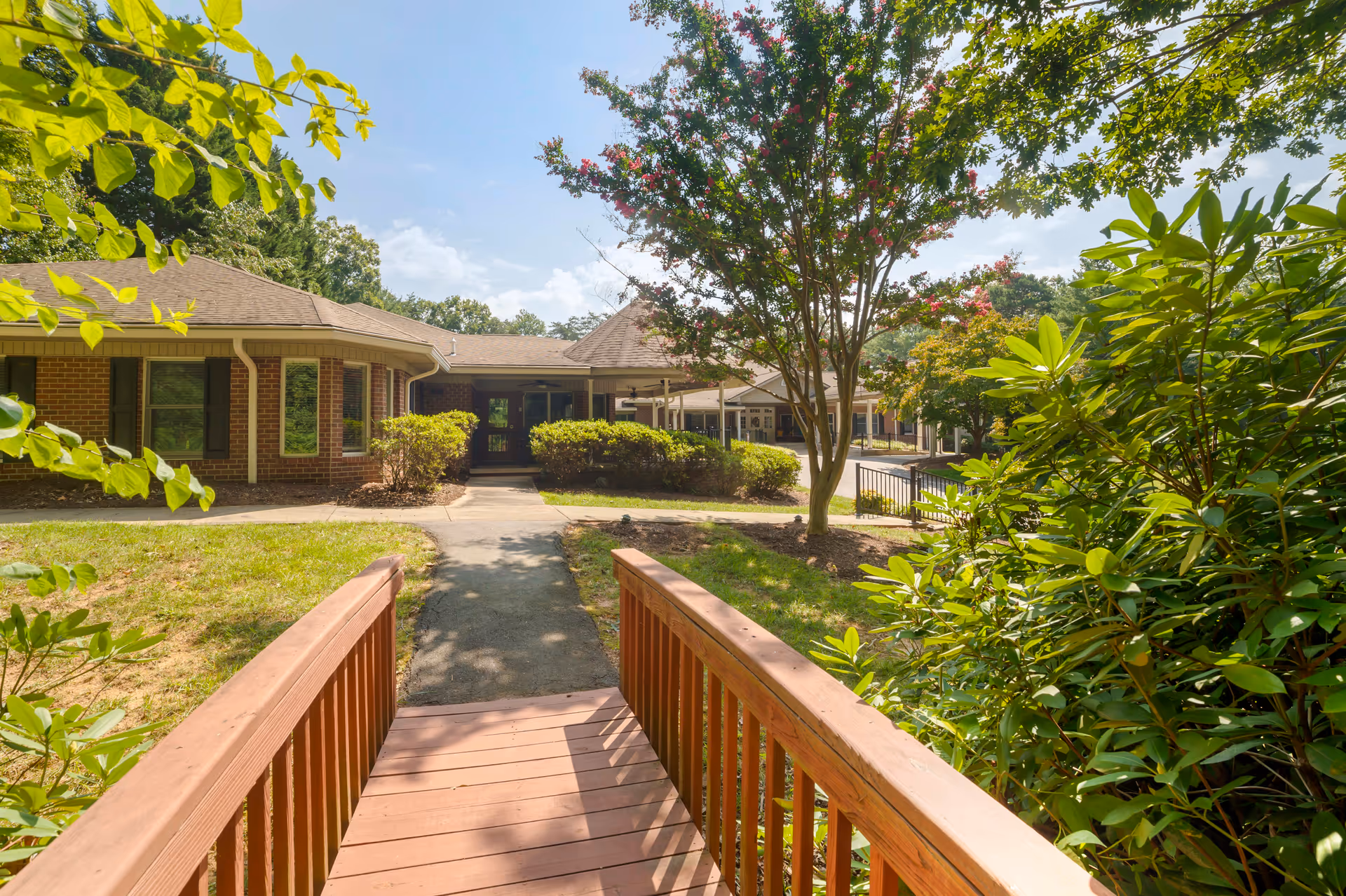 View of the exterior of a senior living facility building with a wooden ramp leading to the entrance. The building is surrounded by green bushes, trees, and a well-maintained lawn under a clear blue sky.