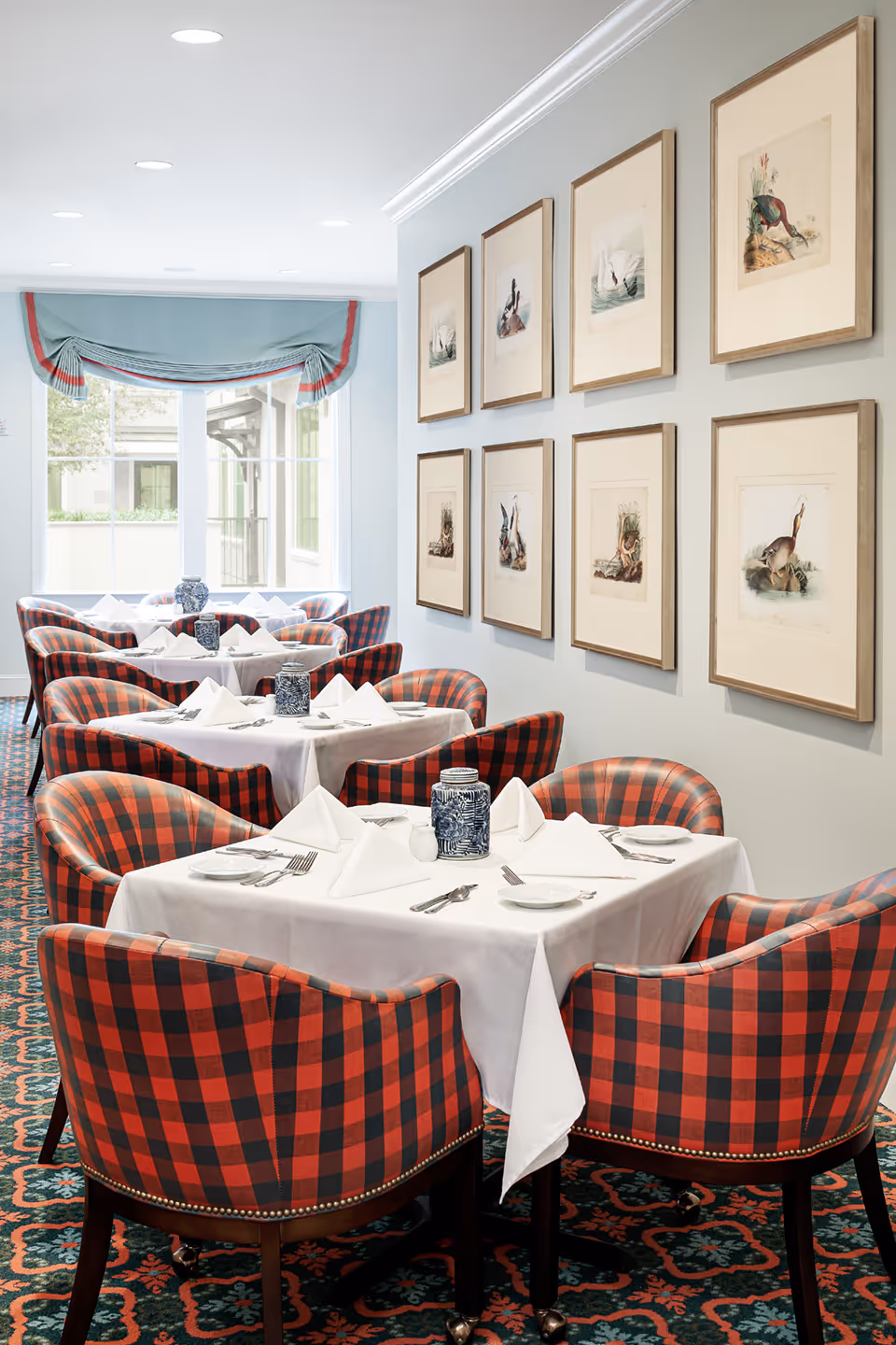 A bright dining room with tables covered in white tablecloths, each set with folded white napkins, plates, and silverware. The chairs around the tables have a red and black checkered pattern. The wall on the right side features a series of framed bird illustrations. A large window with a blue valance with red trim lets in natural light at the far end of the room.