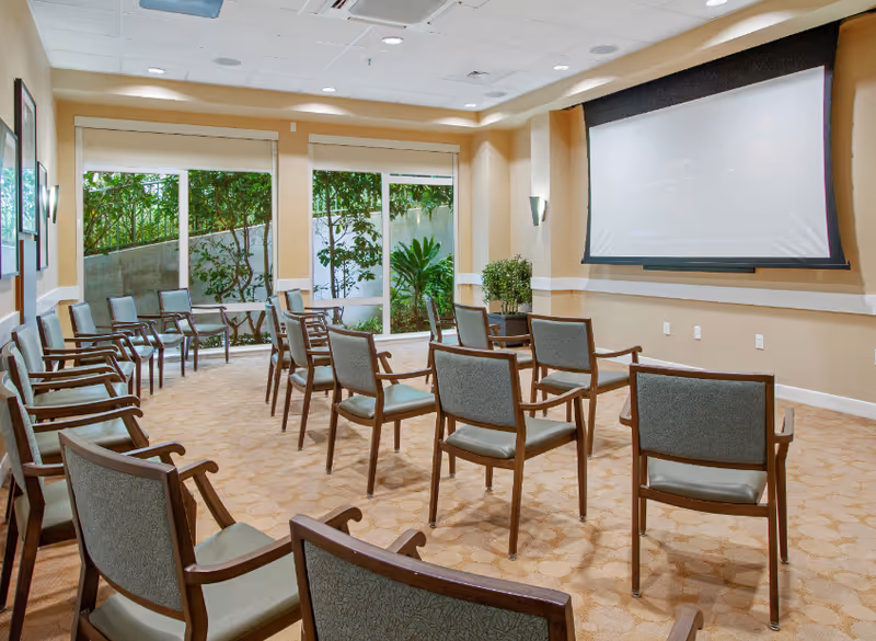 A small auditorium or meeting room with multiple rows of wooden chairs with green cushions arranged facing a large white projection screen on the wall. The room has beige walls, carpeted floor, and large windows showing greenery outside.