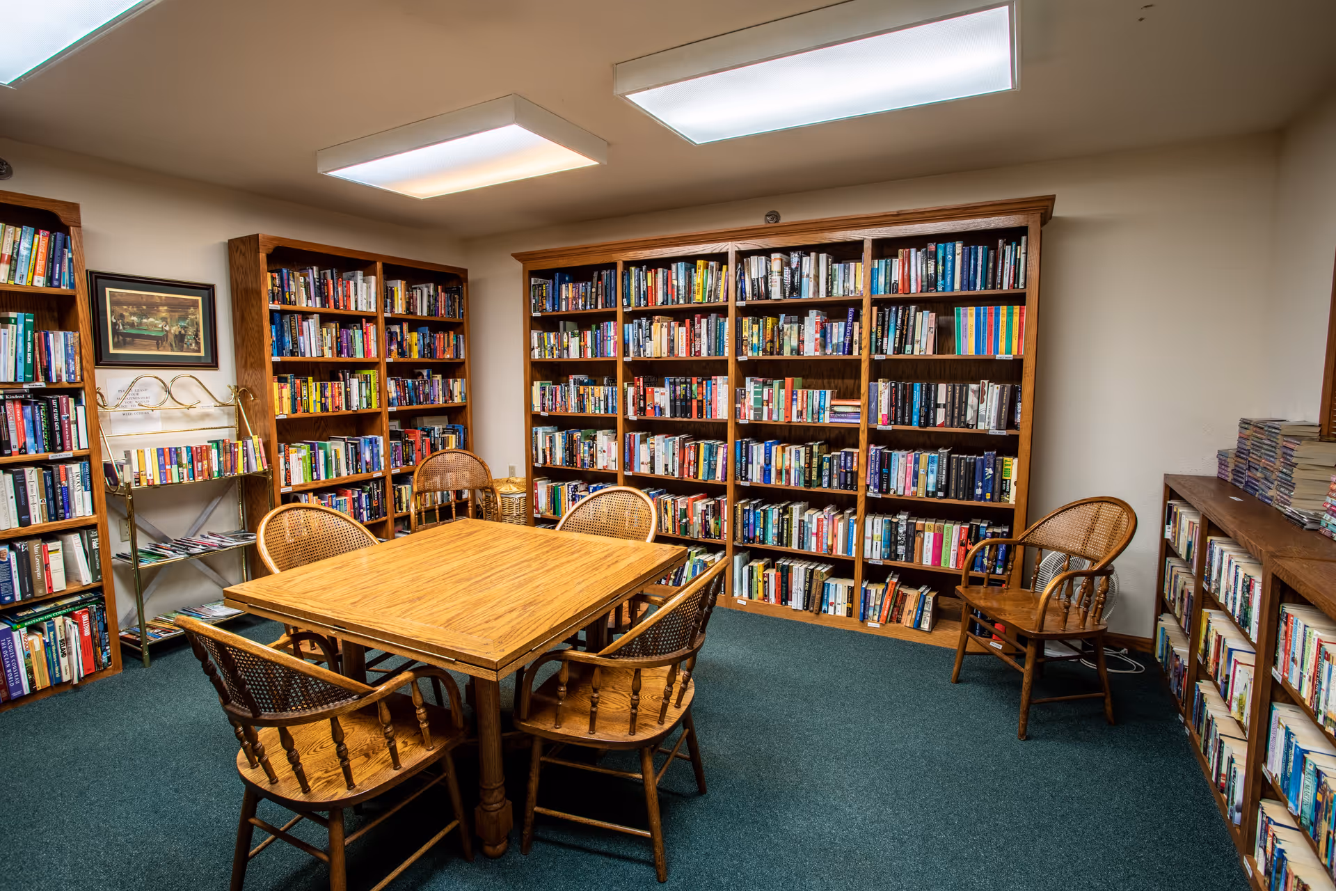 A cozy library room with wooden bookshelves filled with books lining the walls. In the center, there is a wooden table surrounded by six wooden chairs with woven backs. The room has a green carpet and bright overhead lighting.