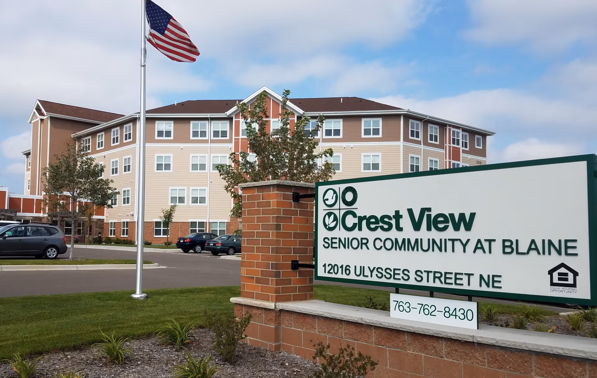 Exterior view of Crest View Senior Community building with a large sign in the foreground displaying the facility name, address, and phone number. An American flag is flying on a flagpole near the building, and several cars are parked in the parking lot.