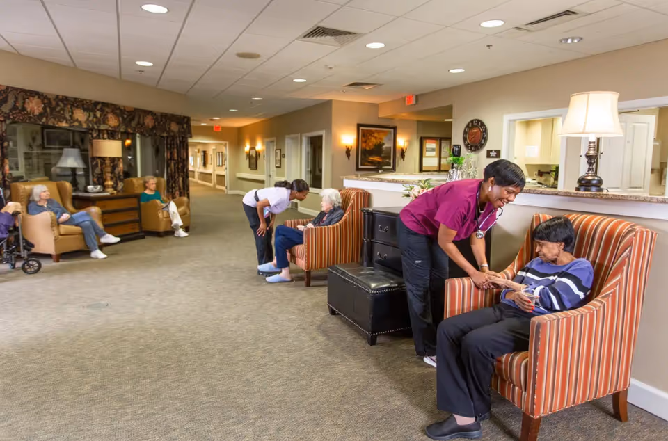 A hallway in an assisted living facility with elderly residents seated in armchairs along the walls. Two caregivers are attending to two elderly women sitting in striped armchairs near a reception desk. Other residents are seated further down the hallway in comfortable chairs. The area is well-lit with ceiling lights and wall sconces, and decorated with framed artwork and floral curtains.